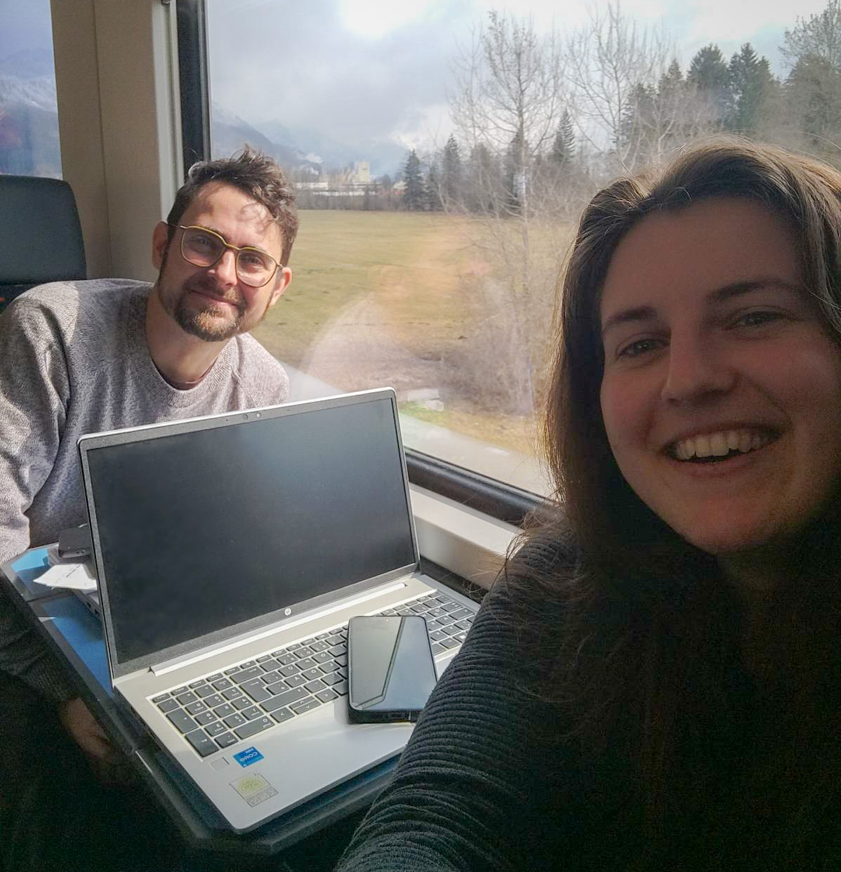 Ein Selfie von zwei Mitarbeiter:innen die im Zug sitzen, am Tisch steht ein Laptop, es handelt sich um eine Dienstreise